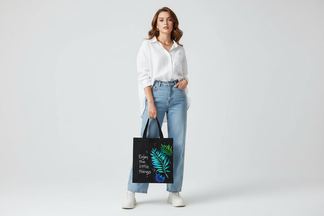 Black tote bag with green leaf design and 'Enjoy the little things' text on a wooden surface
