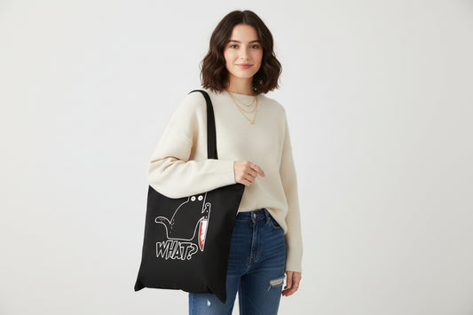 Black tote bag with a cat holding a knife and 'WHAT?' text, held by a person against a neutral background.